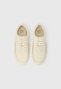 Kreemivärviliste BOSSi tossude paar, millel on paelad, Ortholite sisetallad ja ümarad varbad valgel taustal.