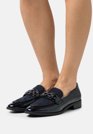 Mocassins en cuir verni noir et en daim avec un détail en chaîne, portés pieds nus sur fond blanc.