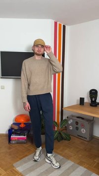 Šviesiai rudas mezgimo megztinis su ribotu apačioje, tamsiai mėlyni siaurėjantys kelnės ir kreminė kepurė. Pilki sportbačiai su žaliais akcentais. Vidaus aplinka su dekoru.