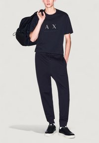 T-shirt en coton noir avec logo "AX", associé à un pantalon de survêtement noir avec ceinture élastique. Des baskets noires complètent la tenue.