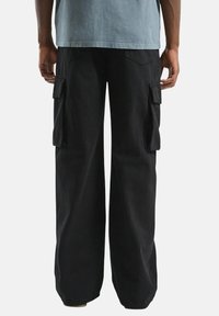 Achteraanzicht van een persoon in zwarte brede cargobroek met klepzakken en een lichtblauw T-shirt.