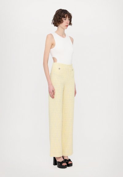 Valkoinen hihaton bodysuit, jossa on sivurei'ät, yhdistettynä korkea vyötäröisiin keltapohjaisiin tweed-laajalahkeisiin housuihin, joissa on kaksi etunappia. Mustat platform-kengät.