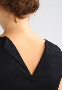 Robe noire en tissu texturé, avec un décolleté en V au dos et une fermeture éclair. Fini lisse avec des motifs subtils.