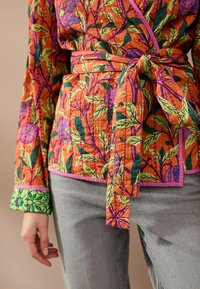 Haut cache-cœur floral coloré orange avec des motifs verts et violets, noué à la taille, présentant un tissu texturé et des poignets contrastants.
