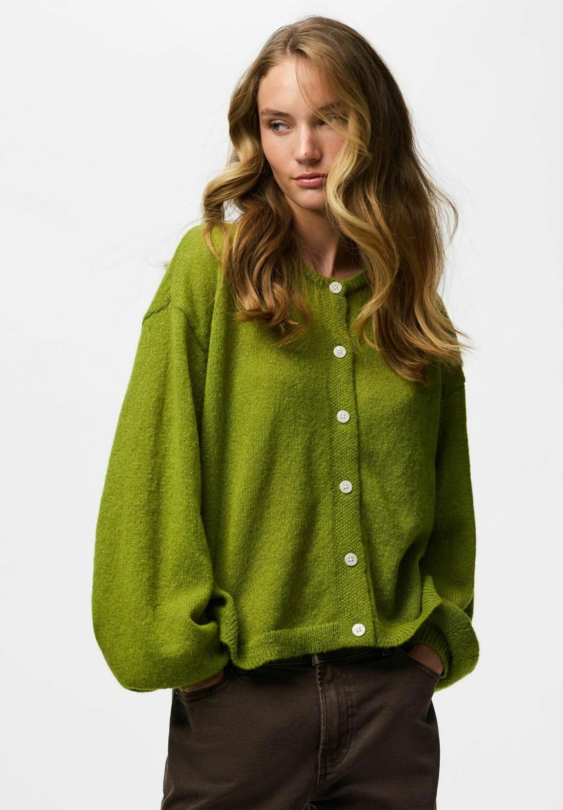 Jeune femme aux longs cheveux ondulés portant un cardigan vert oversize avec des boutons blancs et un pantalon foncé, les mains dans les poches, regardant sur le côté.