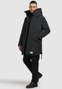 Parka izolată de culoare neagră, cu guler înalt, închidere cu fermoar și capse, multiple buzunare, căptușeală texturată și patch cu logo la margine.