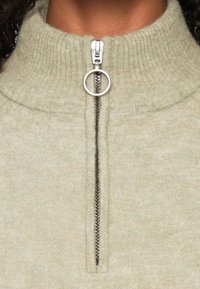 Suéter de color verde claro con cremallera y cuello acanalado. Cuenta con una cremallera plateada con un tirador circular y una tela suave y texturizada.