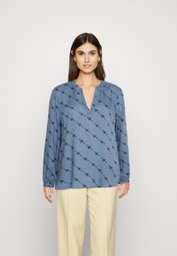 Blusa azul de mangas longas em tecido leve, com decote em V e um padrão floral preto. Combinada com calças de tom claro.