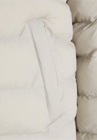 Tæt på puffet jakke i cremefarve, der har quiltede sektioner, synlige sømme og en sidelomme med matchende stofkant.