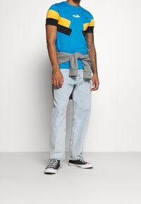 Camiseta de algodón azul con detalles amarillos y negros, combinada con jeans azul claro y zapatillas negras. Sudadera gris atada a la cintura.