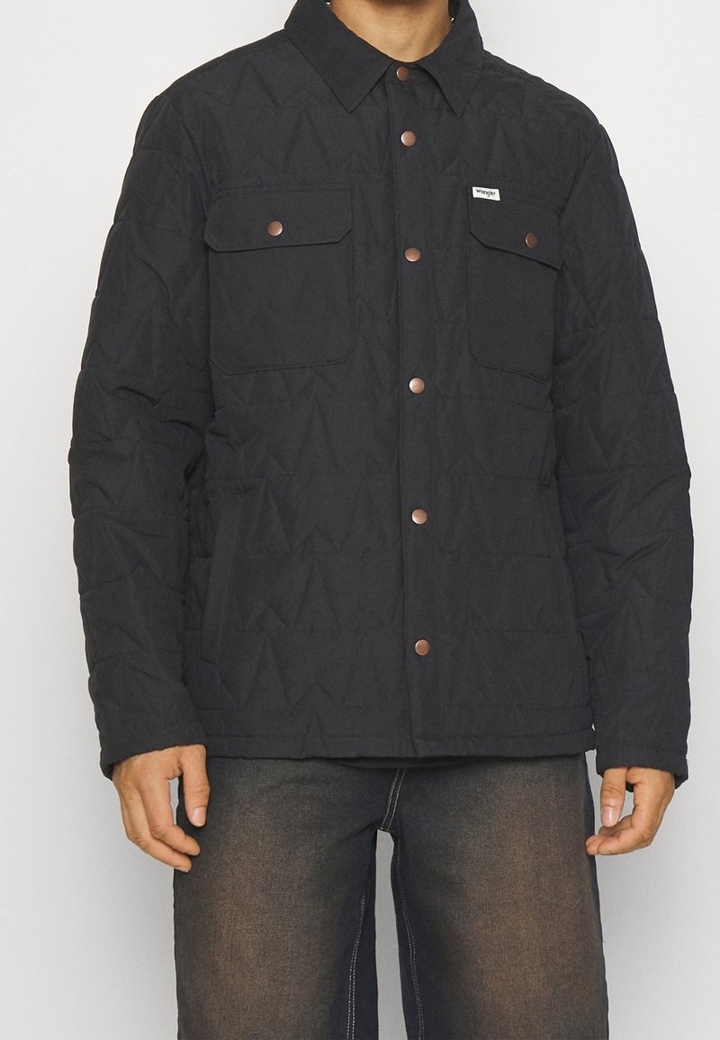 Man wearing a black quilted jacket with copper snap buttons and two chest flap pockets, paired with dark faded jeans.