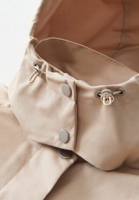 Collo della giacca beige con cappuccio, con accenti in metallo argentato, inclusi bottoni a pressione e cordoncino. Tessuto liscio e textured con sottili righe verticali.