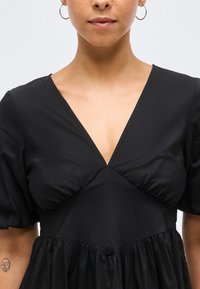 Γυναίκα με μαύρο φόρεμα με λαιμόκοψη σε σχήμα V, φουσκωτά μανίκια και κρίκους σκουλαρίκια, που εμφανίζει μερικώς το πρόσωπο και το πάνω μέρος του κορμού της σε απλό φόντο.