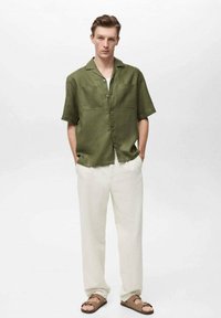 Short-sleeve olive green shirt with two front pockets and buttons, paired with loose-fitting cream trousers and brown sandals.