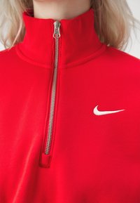 Röd zip-tröja i mjukt material, med hög krage och silverdragkedja, samt en liten vit Nike-logga på bröstet.