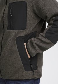 Fleecejack in olijfgroen met zwarte nylon accenten. Voorzien van een ritssluiting op de borst en zijzakken, gestructureerde stof en elastische manchetten.