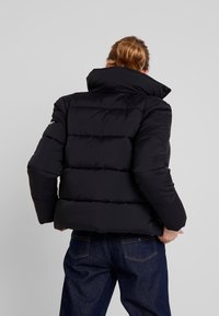 Svart puffjacka med hög krage, med horisontella quiltade paneler. Materialet verkar slätt och lättviktigt. Synlig logotyp på ärmen.