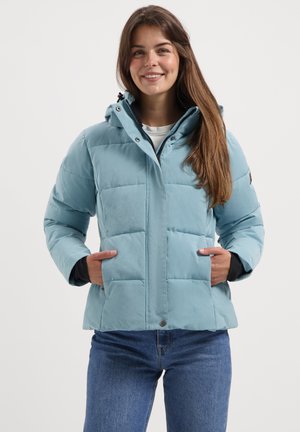 Lachende vrouw met lang bruin haar, gekleed in een lichtblauwe pufferjack met capuchon en een spijkerbroek, met haar handen in de zakken van de jas.