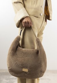 Sac à main en peluche marron avec une forme arrondie et une texture douce. Il dispose d'une courte sangle et d'une petite plaque logo à l'avant.