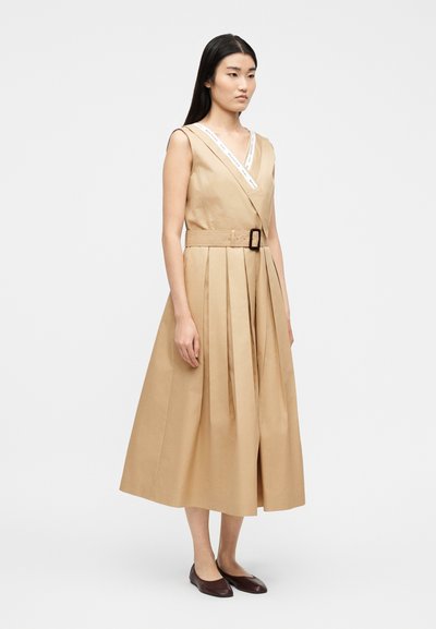 Γυναίκα που φοράει αμάνικο μπεζ μεσαίο φόρεμα με πιέτες και ζώνη στη μέση, συνδυασμένο με σκούρα καφέ επίπεδα παπούτσια, στέκεται μπροστά σε απλό φόντο.