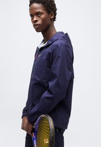 Νεαρός άνδρας με μπλε μπουφάν που κρατά ρακέτα του τένις με κίτρινο σχέδιο χορδών μπροστά σε απλό φωτεινό φόντο.