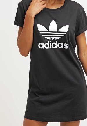 Personne portant une robe T-shirt noire Adidas avec le logo trefoil blanc d'Adidas sur la poitrine, une main près du col.