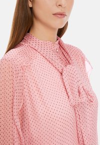 Rosa Bluse aus leichtem Stoff, mit einer Schleife am Hals, vertikalen Rüschen-Details und einem gepunkteten Muster in dunkleren Tönen.