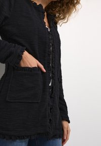 Czarna teksturowana kurtka z falbankowymi brzegami, zapięciami na guziki i przednią kieszenią. Posiada długie rękawy i wygodne dopasowanie.