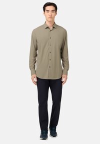 Khaki overhemd met lange mouwen, een button-down kraag en knopen aan de voorkant, gecombineerd met donkere broek en blauwe sneakers, tegen een effen achtergrond.