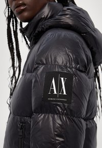 Pufasta jakna u sjajnom crnom najlonu s visokim ovratnikom. Ima „A/X“ logo na rukavu i zatvaranje na zip.