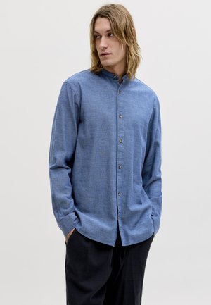 Giovane uomo con capelli fino alle spalle che indossa una camicia blu a maniche lunghe con bottoni e pantaloni neri, che guarda a sinistra con le mani in tasca.