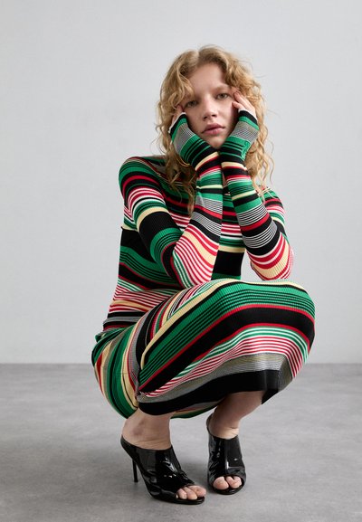 Abito in maglia a coste multicolore con righe orizzontali in verde, rosso e nero; maniche lunghe, con aperture per il pollice; abbinato a sandali neri con tacco.