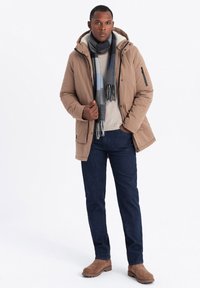 Giubbotto imbottito color sabbia con cappuccio, tasche laterali e dettagli in zip nera, indossato sopra un maglione chiaro, jeans scuri e stivaletti marroni.
