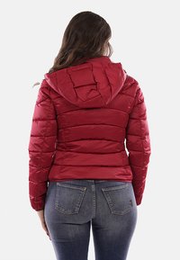 Giacca a vento rossa con cappuccio, design trapuntato e finitura lucida. Presenta polsini elasticizzati e una silhouette aderente, abbinata a jeans blu.