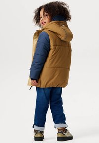 Puffige, zweifarbige Jacke mit braunem Körper und marineblauen Ärmeln. Verfügt über eine Kapuze und Reißverschluss-Taschen. Wird mit hochgekrempelten Jeans und Schuhwerk getragen.