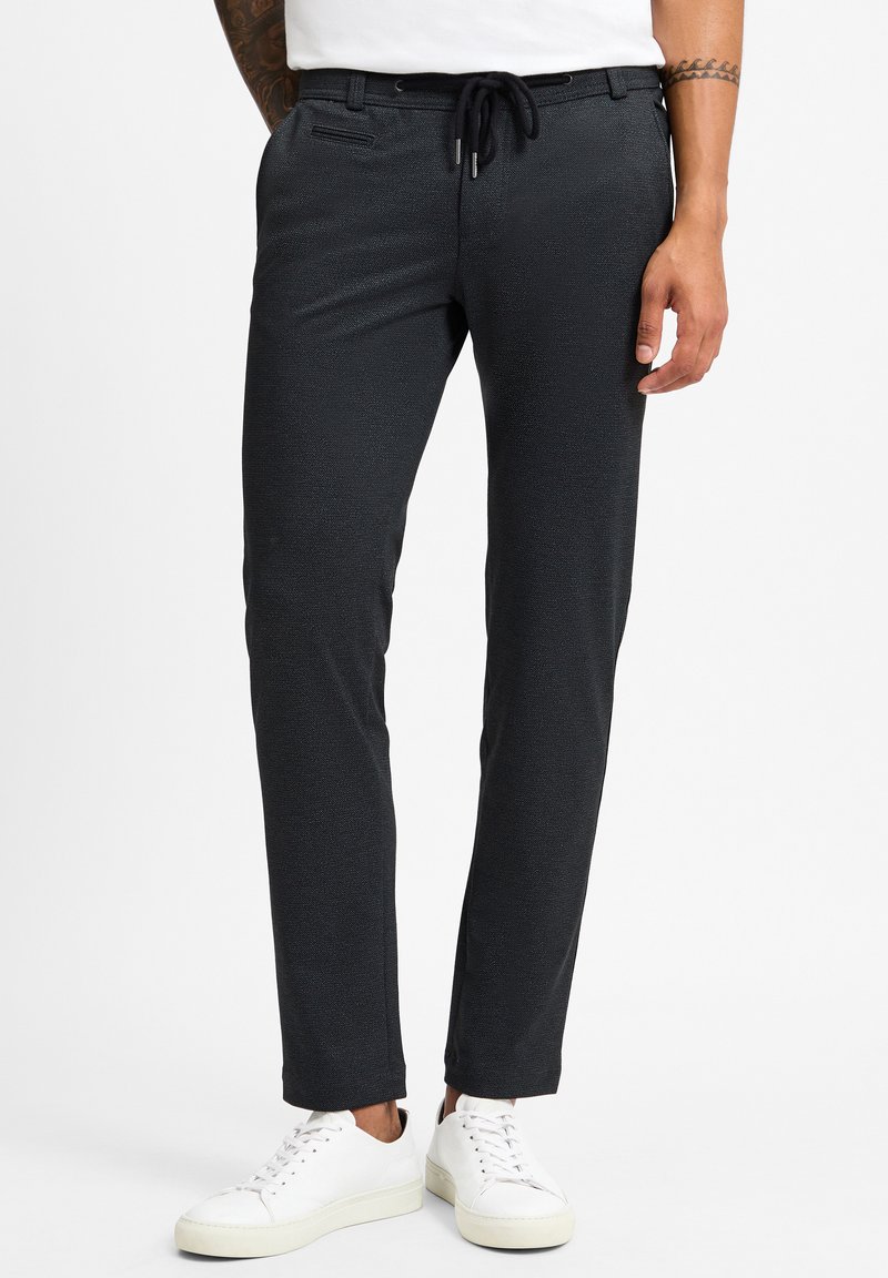 Schwarze, strukturierte Hose mit einem Kordelzugbund und seitlichen Taschen. Kombiniert mit weißen Sneakers und einem schmalen Schnitt.