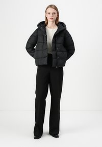 Casaco puffer preto com capuz, apresentando uma textura acolchoada e um fecho frontal. Usado sobre um suéter leve e calças pretas.