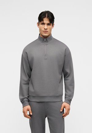 Άνδρας που φοράει γκρι πουλόβερ με μακριά μανίκια και φερμουάρ στο μισό στόμιο και ταιριαστό γκρι παντελόνι, στέκεται μπροστά σε απλό φόντο.