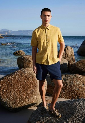Muž v horčicovej krátkorukávovej košeli na zapínanie, tmavomodrých šortkách a hnedých lodných topánkach stojaci na skalách pri oceáne pod jasnou oblohou.