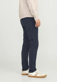 Marineblaue Hose in Slim Fit-Design, aus glattem Stoff. Getragen mit einem beigen Langarmshirt und weißen Sneakers mit tanfarbenen Akzenten.