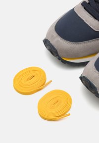 Sneaker in suede grigio e mesh blu, con un accento giallo sulla suola. Due lacci gialli arrotolati posizionati accanto alla scarpa.