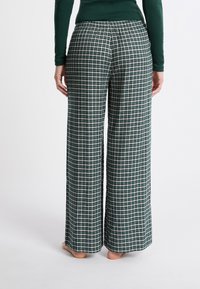 Persoon in losse, hoog geheven groen-wit geblokte broek en een donkergroene langemouwen top, staat blootsvoets op een witte vloer.