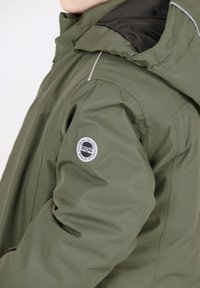 Giacca esterna verde con cappuccio, dotata di colletto nero, dettagli riflettenti e un patch circolare con logo sulla manica sinistra.