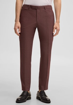 Mężczyzna ubrany w dopasowane, brązowe eleganckie spodnie, w połączeniu z czarnymi eleganckimi butami na sznurowadła, stojący na tle jednolitego szarego tła.