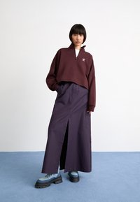 Sudadera corta burdeos con cremallera de cuarto y logo bordado, combinada con una falda larga plisada de color púrpura oscuro y botas azules con cordones.