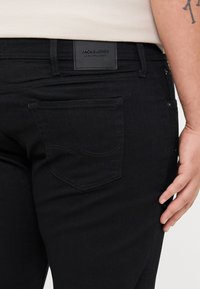Crne traperice od denima s stražnjim zakrpatom s oznakom "JACK & JONES." Značajke uključuju zakrivljeno šav detalje na desnom džepu i uski kroj.