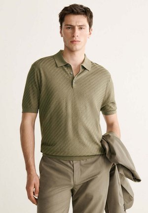 Jonge man die een gestructureerd olijfgroen poloshirt met korte mouwen en bijpassende broek draagt, en in zijn linkerhand een opgevouwen jas vasthoudt.