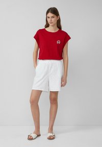 Rode top met korte mouwen met een kleine krabprint, gecombineerd met witte oogjeskorte broek met een trekkoord taille. Witte sandalen maken de look af.