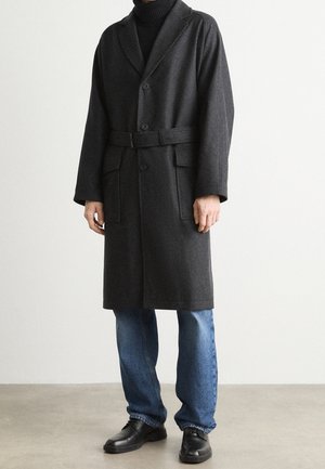 Homme portant un manteau en laine gris foncé ceinturé par-dessus un col roulé noir, un jean bleu et des chaussures en cuir noir, debout contre un mur blanc.