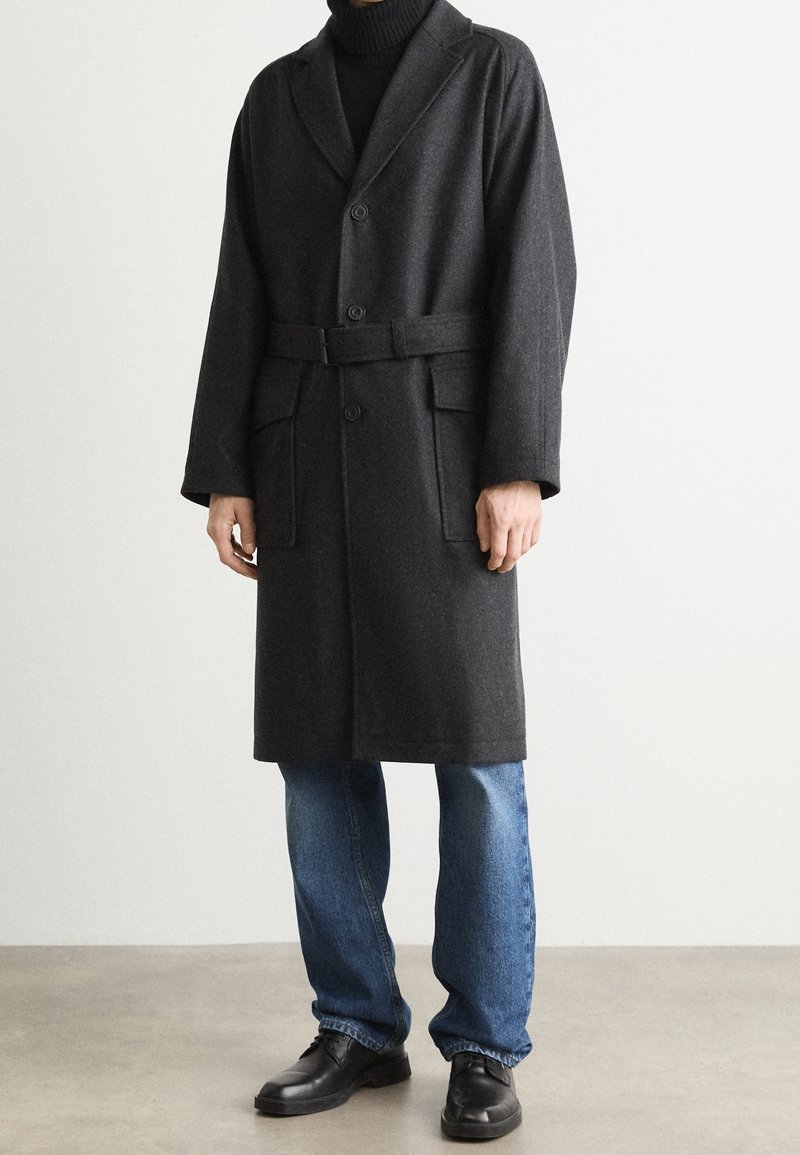 Homme portant un manteau en laine gris foncé ceinturé par-dessus un col roulé noir, un jean bleu et des chaussures en cuir noir, debout contre un mur blanc.